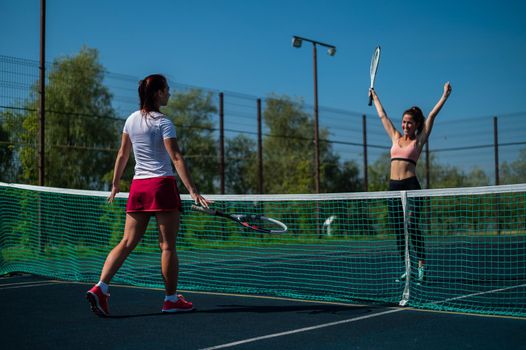 Two athletic young women play tennis on an outdoor court on a hot summer day. The girl is happy with the victory over her rival
