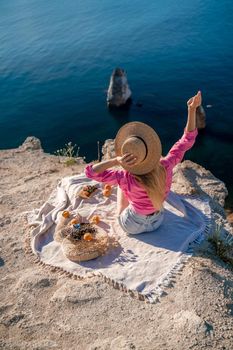 photo of a beautiful woman with long blond hair in a pink shirt and denim shorts and a hat having a picnic on a hill overlooking the sea.