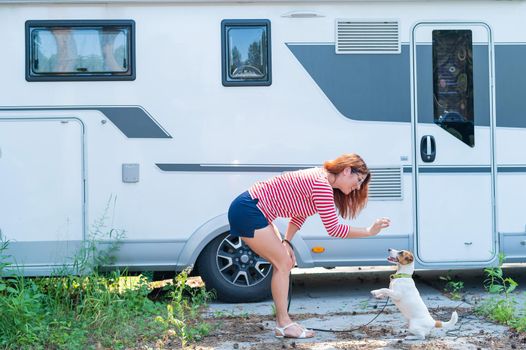 Beautiful caucasian woman is training a dog next to the motorhome. Travel with your pet near the mobile home on wheels. Redhead Female Trailer Driver plays with Jack Russell Terrier