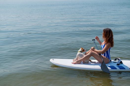 A woman is riding a sup surfboard with a dog on the lake. The girl goes in for water sports with her pet