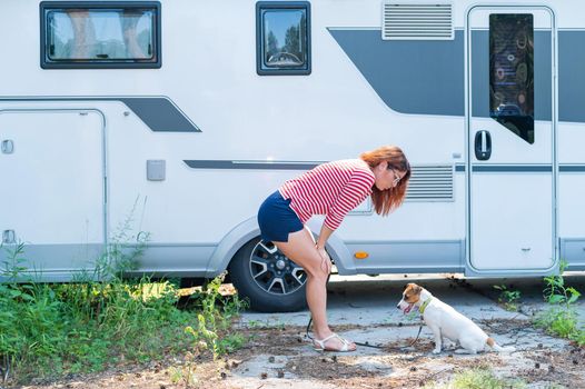 Beautiful caucasian woman is training a dog next to the motorhome. Travel with your pet in a mobile RV. Red-haired girl playing with Jack Russell Terrier. Female trailer driver