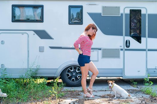 Beautiful caucasian woman is training a dog next to the motorhome. Travel with your pet in a mobile RV. Red-haired girl playing with Jack Russell Terrier. Female trailer driver