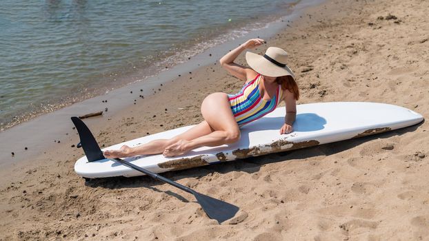 A woman in a swimsuit and a hat is posing lying on a sup board on the beach