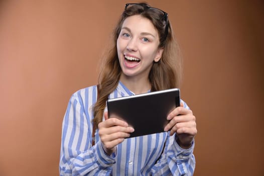 Portrait of a cheerful charming girl holding a digital tablet with a toothy smile, wearing a shirt and glasses, studio shot.