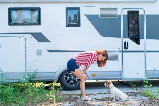 Beautiful caucasian woman is training a dog next to the motorhome. Travel with your pet in a mobile RV. Red-haired girl playing with Jack Russell Terrier. Female trailer driver