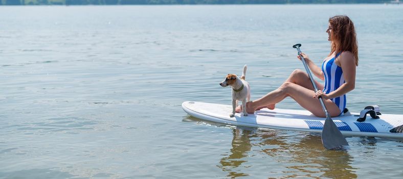 A woman is riding a sup surfboard with a dog on the lake. The girl goes in for water sports with her pet