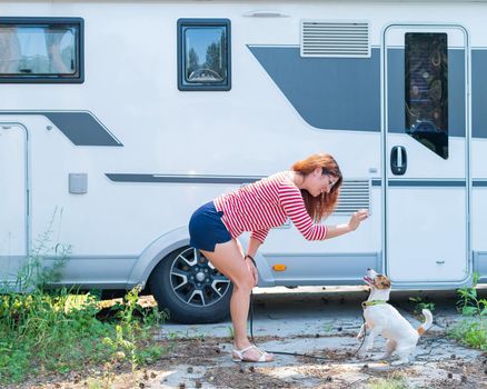 Beautiful caucasian woman is training a dog next to the motorhome. Travel with your pet in a mobile RV. Red-haired girl playing with Jack Russell Terrier. Female trailer driver