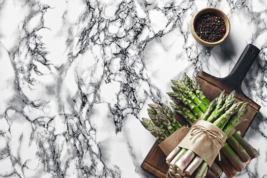 Bunch of an edible, fresh sprigs of asparagus on a wooden board, marble background. Green vegetables with seasonings, top view. Healthy eating. Spring harvest, agricultural farming concept.