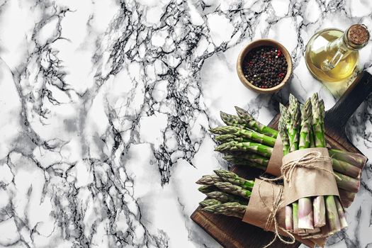 Bunch of an edible, fresh sprouts of asparagus on a wooden board, marble background. Green vegetables with olive oil and seasonings, top view. Healthy food. Spring harvest, agricultural farming concept.