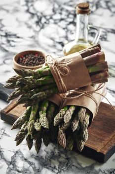 Bunch of an edible, green stems of asparagus on a wooden board, marble background. Fresh vegetables with olive oil and seasonings, top view. Healthy eating. Spring harvest, agricultural farming concept.