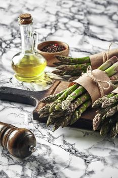 Bunch of an edible, raw sprouts of asparagus on a wooden board, marble background. Fresh, green vegetables with olive oil and seasonings. Healthy eating. Spring harvest, agricultural farming concept.
