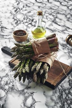 Bunch of an edible, green sprigs of asparagus on a wooden board, marble background. Fresh vegetables with olive oil and seasonings, top view. Healthy eating. Spring harvest, agricultural farming concept.