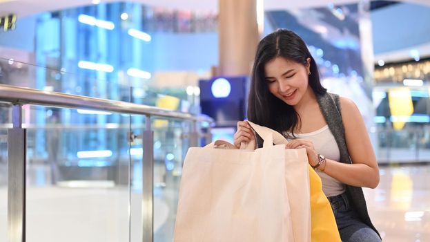 Happy young asian woman sitting on bench in shopping mall with paper bags.