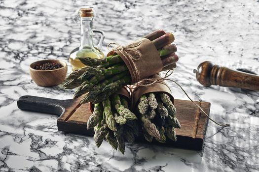 Bunch of an edible, green sprouts of asparagus on a wooden board, marble background. Fresh vegetables with olive oil and seasonings, top view. Healthy food. Spring harvest, agricultural farming concept.