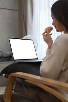 Side view of young woman in warm woolen sweater eating donut and using laptop in cozy living room.