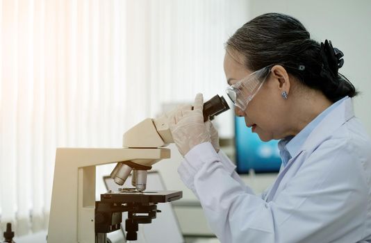 Medical Development Laboratory: Female Scientist Looking Under Microscope, Analyzes Petri Dish Sample. In Background Big Pharmaceutical Lab with Specialists Conducting Medicine, Biotechnology Research.