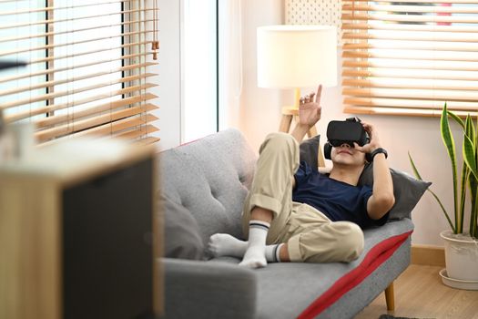 Young man in casual clothes lying on couch and watching movie or play video game on virtual reality headset.