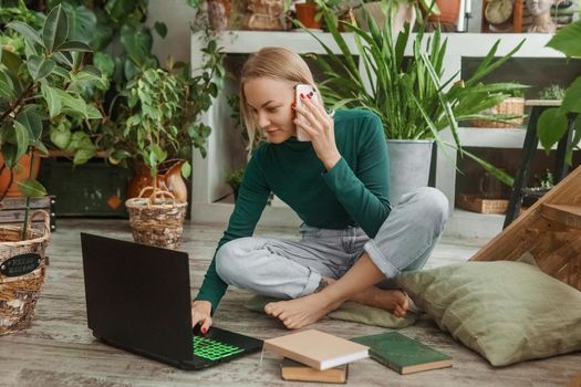 A blonde woman in a room with a lot of green indoor plants is working on a laptop. The concept of biophysical design in the interior. Work from home, work as a freelancer.