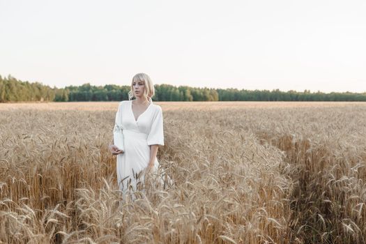 A blonde woman in a long white dress walks in a wheat field. The concept of a wedding and walking in nature.