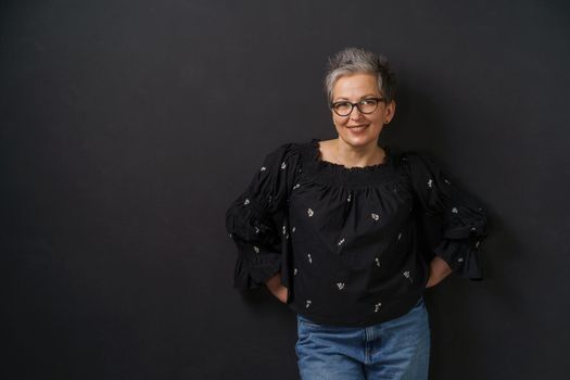 Gorgeous mature grey haired businesswoman with grey hair leaned on wall isolated on black background. Elegant woman with short hair. Image of successful mature woman. Aged beauty.