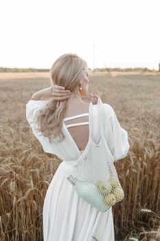 A blonde woman in a long white dress walks in a wheat field. The concept of a wedding and walking in nature.