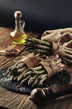 Bunches of an edible, organic sprigs of asparagus on a stone slate, wooden table. Fresh, green vegetables with olive oil and seasonings, kitchen pestle. Healthy eating. Fall harvest, agricultural farming concept.
