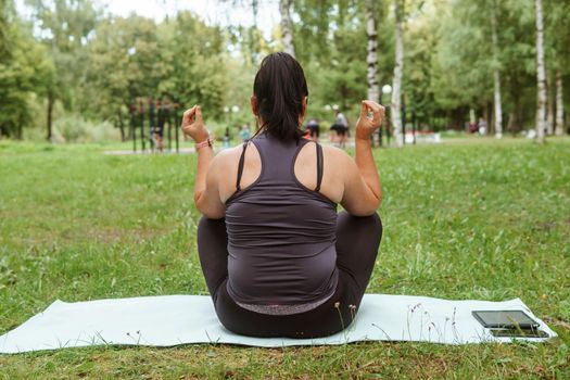 A charming brunette woman plus-size body positive practices sports in nature. Woman does yoga and meditation in the park on a sports mat