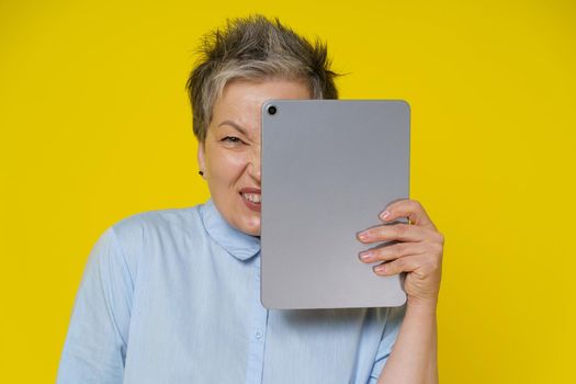 Online troll mature woman hide behind digital tablet with evil facial expression isolated on yellow background. Modern senior woman post bad comments online and happy about it. Hate, bulling concept.