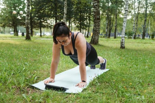 A charming brunette woman plus-size body positive practices sports in nature. Woman does yoga in the park on a sports mat. On a yoga mat in the dog pose.