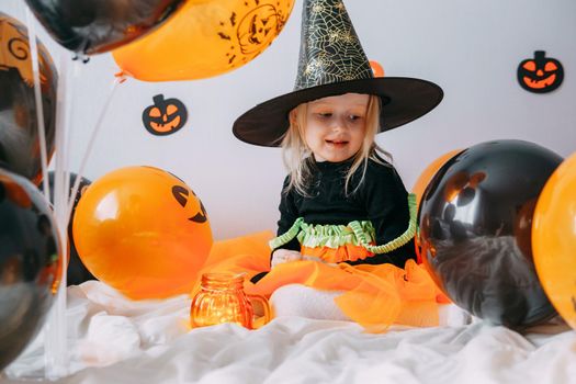 Children's Halloween - a girl in a witch hat and a carnival costume with airy orange and black balloons at home. Ready to celebrate Halloween.