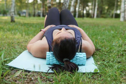 A charming brunette woman plus-size body positive practices sports in nature. Woman is engaged with a massage roller for the body in the park on a sports mat.