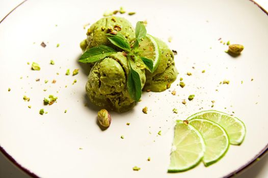 Selective focus on scoops of pistachio sorbet ice cream with sprinkled nuts and green matcha powder on white plate, garnished with fresh juicy lime slices. Raw vegan, dairy free refreshing dessert