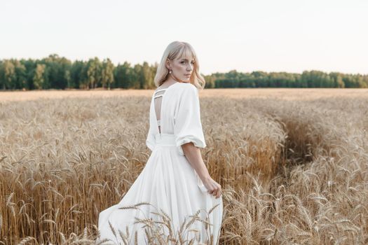 A blonde woman in a long white dress walks in a wheat field. The concept of a wedding and walking in nature.