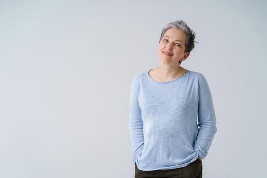 Happy dreamy mature grey haired woman 50s posing tenderly looking at camera with hands in pockets, copy space on left isolated on white background. Mature people healthcare.