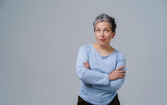Grey haired mature woman standing with arms folded and wondering face expression in blue blouse copy space on left isolated on white background. Healthcare concept. Aged beauty concept. Copy space.