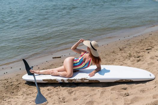 A woman in a swimsuit and a hat is posing lying on a sup board on the beach