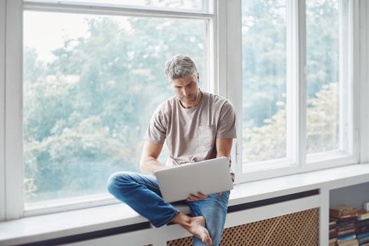 man using a laptop sitting on the windowsill in the living room. photo with a copy-space.