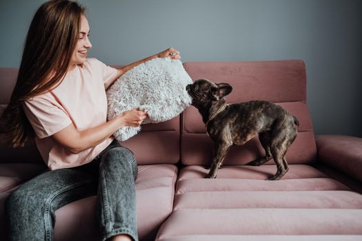 French Bulldog Biting Pillow on Pink Sofa at Home While Cheerful Woman Trying to Protect Her
