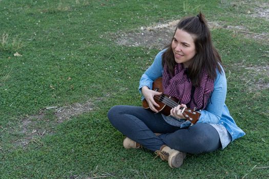 young brunette girl with long hair playing ukulele in the field on the grass