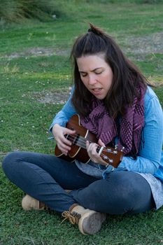 young brunette girl with long hair playing ukulele in the field on the grass