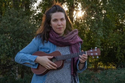 young brunette girl with long hair playing ukulele in the field on the grass