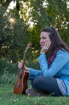 young brunette girl with long hair playing ukulele in the field on the grass