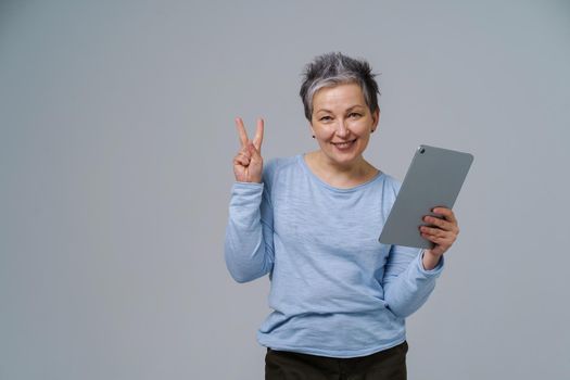 Gesturing V or victory mature grey haired woman in 50s holding digital tablet working or shopping online, checking on social media. Pretty woman in blue blouse isolated on white background.