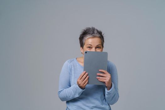 Shy mature woman hide holding digital tablet working or shopping online, checking on social media. Pretty grey haired woman in blue blouse isolated on white background.
