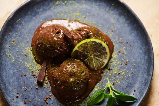 Flat lay. Food still life. Raw vegan chocolate ice cream sorbet balls, garnished with sprinkled green matcha powder, slice of juicy lime and lemon basil leaves on dark blue plate, on marble background