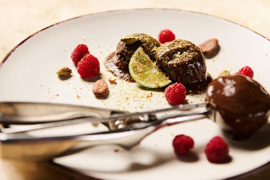 Selective focus on frozen balls of chocolate ice cream on white plate garnished with a slice of juicy lime, pistachios, raspberries and cocoa beans and metal scoop for sorbet on the blurred background