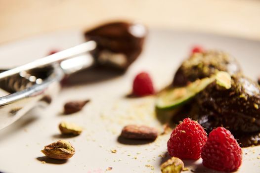 Selective focus on ripe raspberries on a white plate with chocolate ice cream, garnished with a slice of juicy lime, pistachios and cocoa beans and metal scoop for sorbet on the burred background