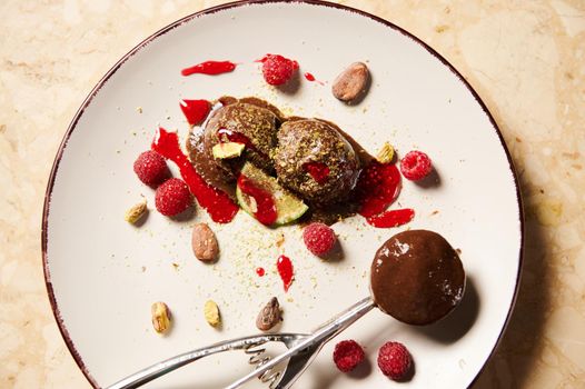 Flat lay of a metal scoop for sorbet, with homemade, dairy free chocolate ice cream, garnished with grated pistachios, raspberries, cocoa beans and berry jam on a white plate. Food still life