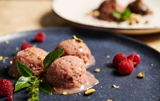 Close-up. Scoops of raspberry ice cream, sorbet with sprinkled pistachio, raspberries and lemon basil on dark blue plate. Healthy raw vegan, refreshing dessert. Summer food still life. Selective focus