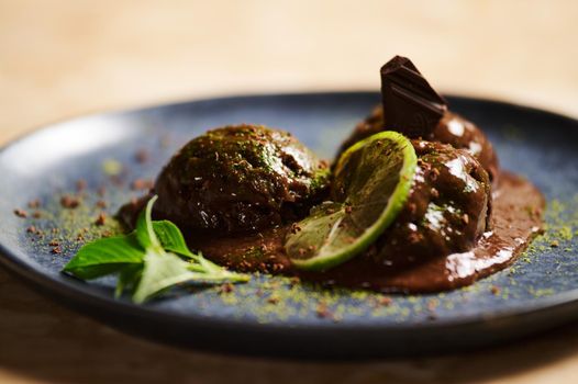 Selective focus. Raw vegan chocolate ice cream sorbet scoops, garnished with sprinkled green matcha powder, slice of juicy lime and lemon basil leaves on dark blue plate, on a marble table background